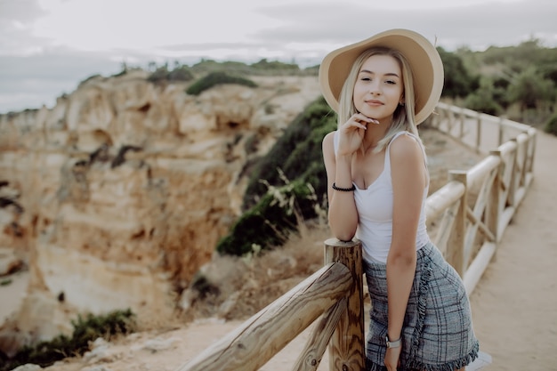 Joven rubia con sombrero y lindo vestido de verano de pie sobre la piedra