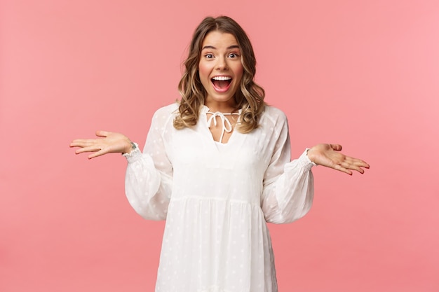 La joven rubia sorprendida y feliz extendió las manos hacia los lados sonriendo, de pie en la pared rosa afortunada y optimista.