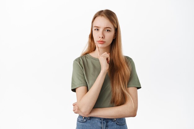 La joven rubia sospechosa tiene dudas, pensando, luciendo seria y pensativa al frente, tomando su decisión, de pie pensativa contra la pared blanca