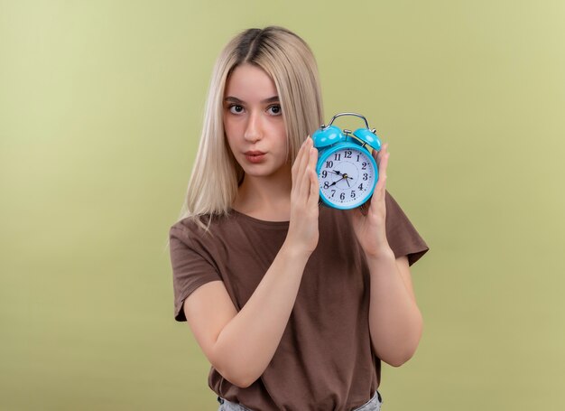 Joven rubia sosteniendo el despertador mirando en la pared verde aislada con espacio de copia