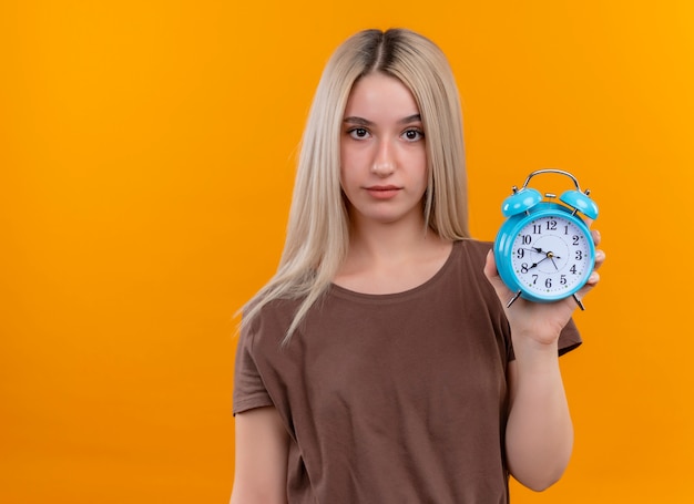 Joven rubia sosteniendo el despertador en la pared naranja aislada con espacio de copia