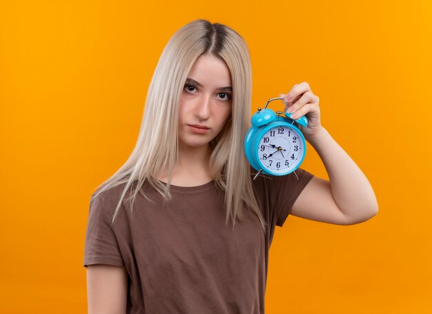 Joven rubia sosteniendo el despertador en la pared naranja aislada