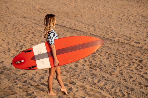 Joven rubia surfista caminando con su tabla de surf en la playa.