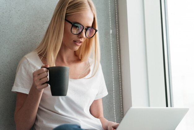 Joven rubia usando laptop y bebiendo té en casa