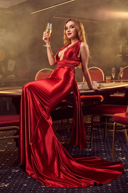 Una joven rubia con un vestido largo de satén rojo, con una copa de champán en la mano, posa sentada en una mesa de póquer en un casino de lujo. Pasión, cartas, fichas, alcohol, ganar, apostar - es una e femenina