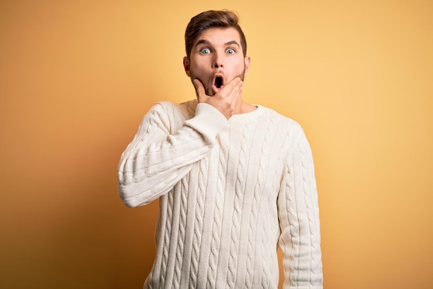 Joven rubio con barba y ojos azules con suéter blanco sobre fondo amarillo Fascinado con sorpresa de incredulidad y expresión asombrada con las manos en la barbilla