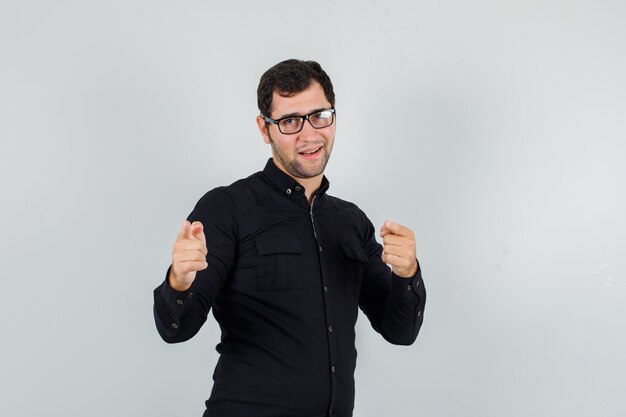 Joven señalando con el dedo a la cámara en camisa negra, gafas y mirando alegre