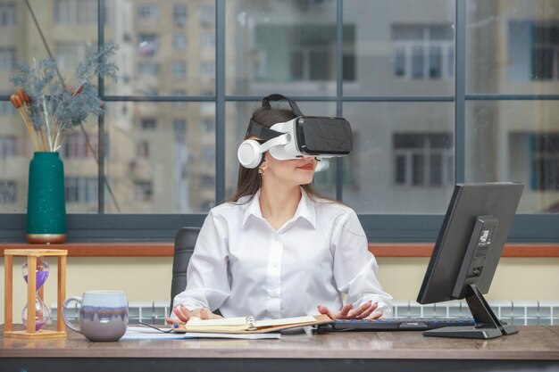 Joven sentada en el escritorio y usando gafas VR mientras mira a un lado Foto de alta calidad