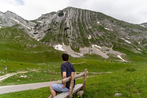 Joven sentado en un banco de madera y disfrutando de la vista de las montañas en un campo de hierba