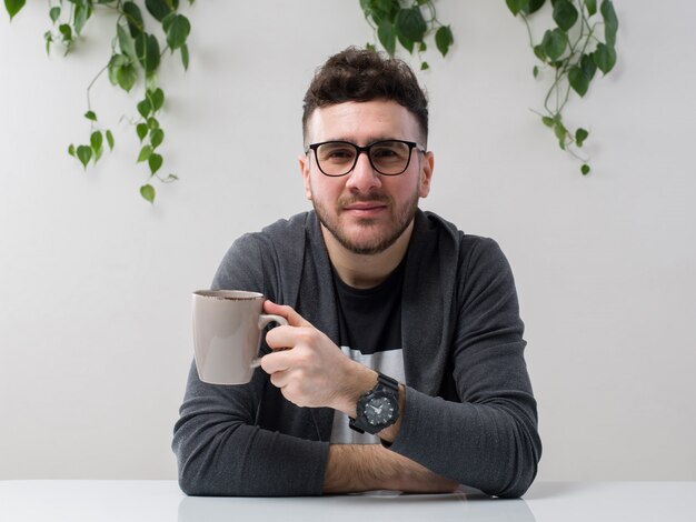 Joven sentado en gafas relojes chaqueta gris sosteniendo su taza de café junto con planta en blanco