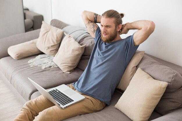 Joven sentado en un gran sofá gris con una laptop y jodiendo los ojos en casa