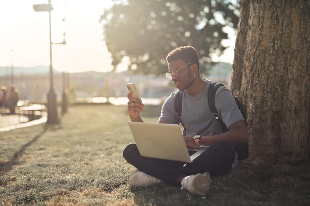 joven sentado en un parque con una computadora y un teléfono inteligente