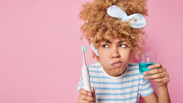 Una joven seria de cabello rizado, enfocada a un lado, usa enjuague bucal y un cepillo de dientes eléctrico limpia los dientes después de despertarse, usa una camiseta a rayas aislada sobre un espacio vacío de fondo rosado para su texto