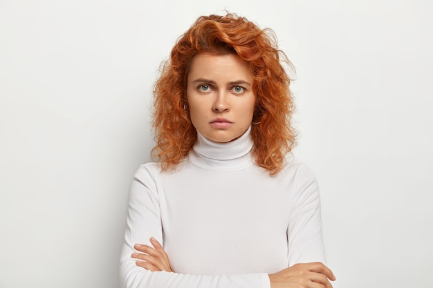 Joven seria con cabello rojo rizado, insatisfecha con algo, mira con enojo, mantiene las manos cruzadas, usa cuello alto blanco informal, ofendida con una pregunta tonta, posa sola en interiores.