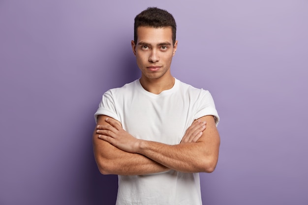 Un joven serio auto asertivo mantiene los brazos musculosos cruzados sobre el pecho, mira directamente, mira atentamente algo, vestido con una camiseta blanca informal, se ve confiado. Estoy listo para escucharte