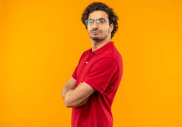 Joven serio en camisa roja con gafas ópticas se encuentra de lado y parece aislado en la pared naranja