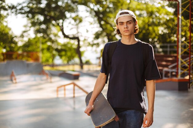 Un joven serio con camiseta negra y gorra blanca mirando cuidadosamente a la cámara mientras sostenía una patineta en la mano con un moderno parque de patinaje en el fondo