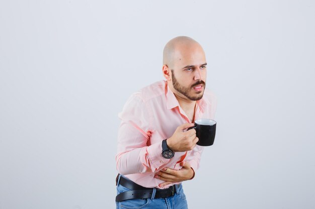 Joven sintiéndose mal después de beber en camisa rosa, jeans y luciendo preocupado. vista frontal.