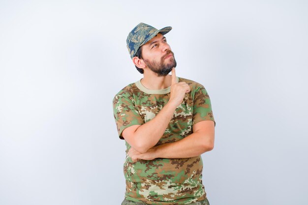 El joven soldado está pensando mirando hacia arriba y sosteniendo el dedo índice debajo de la barbilla sobre fondo blanco.