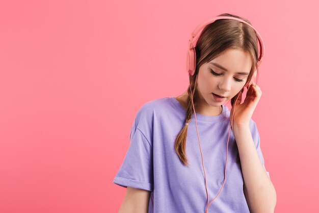 Joven soñadora con dos trenzas en camiseta lila escuchando atentamente música en auriculares mientras pasa tiempo sobre fondo rosa