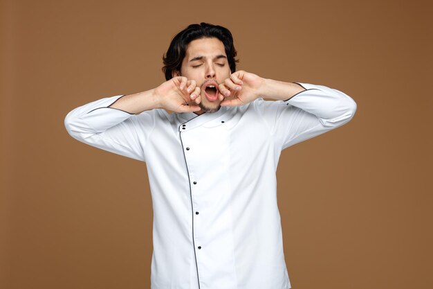 Un joven y soñoliento chef uniformado manteniendo las manos cerca de la boca estirándose y bostezando con los ojos cerrados aislado en un fondo marrón