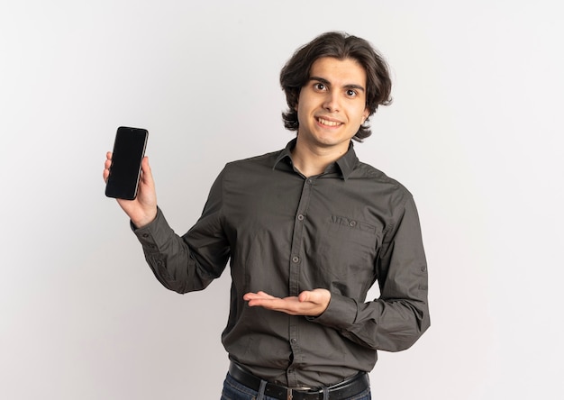 Joven sonriente apuesto hombre caucásico sostiene y apunta al teléfono aislado sobre fondo blanco con espacio de copia