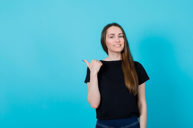 Una joven sonriente apunta hacia atrás con el pulgar en el fondo azul