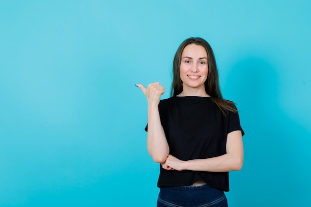 Una joven sonriente apunta a la izquierda con el pulgar en el fondo azul