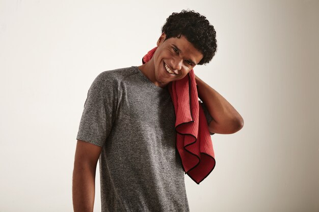 Joven sonriente atleta afroamericano de pelo rizado oscuro con camiseta técnica gris secándose el cuello con una toalla de microfibra waffle roja sobre blanco