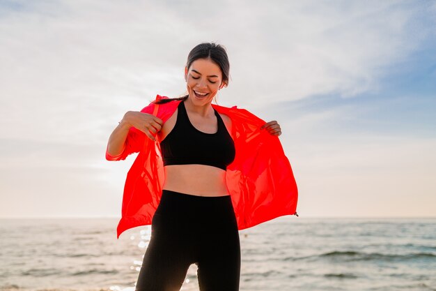 Joven sonriente atractiva mujer delgada haciendo deporte en el amanecer de la mañana bailando en la playa del mar en ropa deportiva, estilo de vida saludable, escuchando música en auriculares, vistiendo chaqueta cortavientos rosa, divirtiéndose