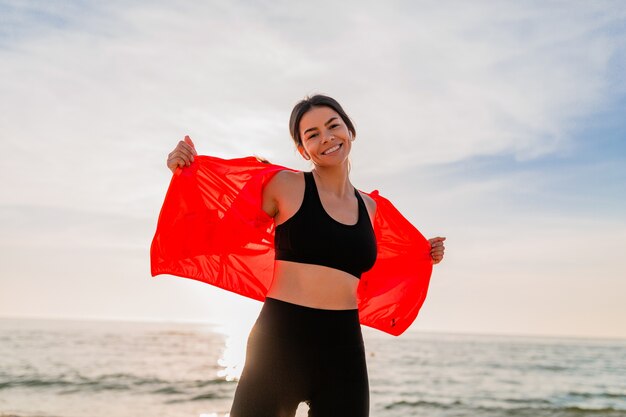 Joven sonriente atractiva mujer delgada haciendo deporte en el amanecer de la mañana bailando en la playa del mar en ropa deportiva, estilo de vida saludable, escuchando música en auriculares, vistiendo chaqueta cortavientos rosa, divirtiéndose