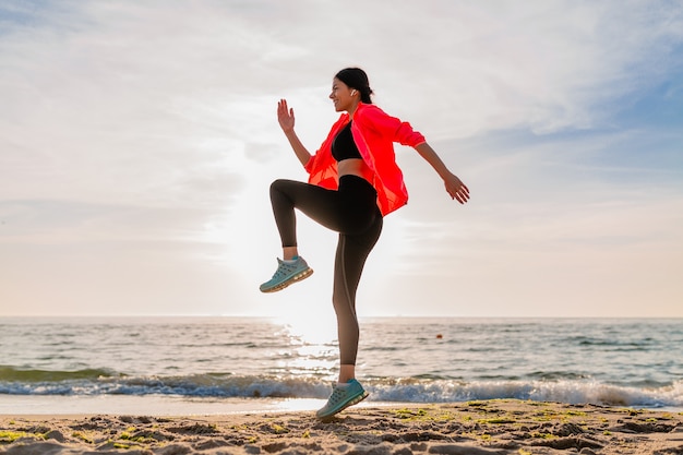 Joven sonriente atractiva mujer delgada haciendo deporte en el amanecer de la mañana saltando en la playa del mar en ropa deportiva, estilo de vida saludable, escuchando música en auriculares, vistiendo chaqueta cortavientos rosa, divirtiéndose