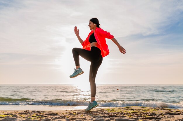 Joven sonriente atractiva mujer delgada haciendo deporte en el amanecer de la mañana saltando en la playa del mar en ropa deportiva, estilo de vida saludable, escuchando música en auriculares, vistiendo chaqueta cortavientos rosa, divirtiéndose