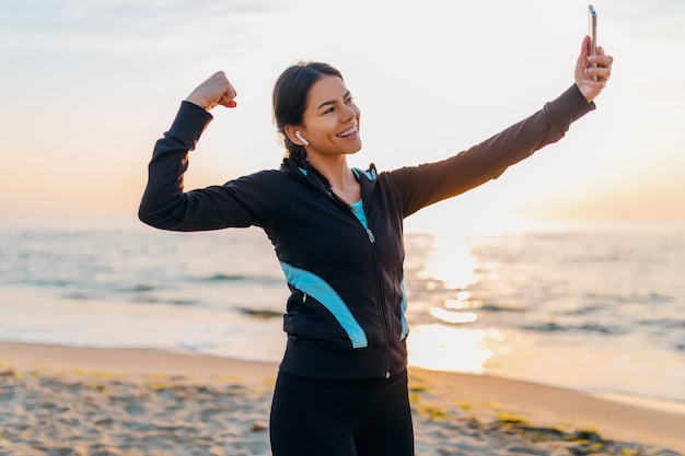 Joven sonriente atractiva mujer delgada haciendo ejercicios deportivos en la playa del amanecer de la mañana en ropa deportiva, estilo de vida saludable, escuchando música en auriculares, haciendo que la foto selfie en el teléfono se vea fuerte