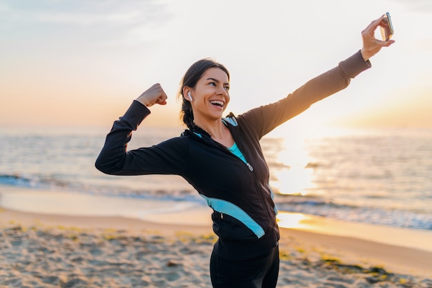 Joven sonriente atractiva mujer delgada haciendo ejercicios deportivos en la playa del amanecer de la mañana en ropa deportiva, estilo de vida saludable, escuchando música en auriculares, haciendo que la foto selfie en el teléfono se vea fuerte