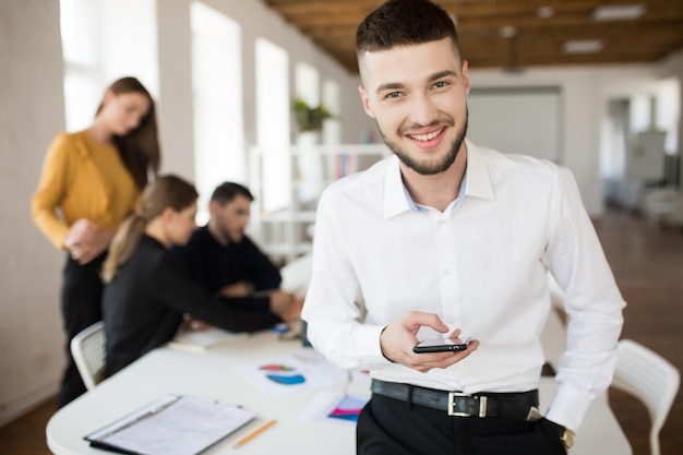 Foto gratuita joven sonriente con barba en camisa blanca mirando felizmente a la cámara sosteniendo el celular en la mano mientras pasa tiempo en la oficina con colegas en el fondo