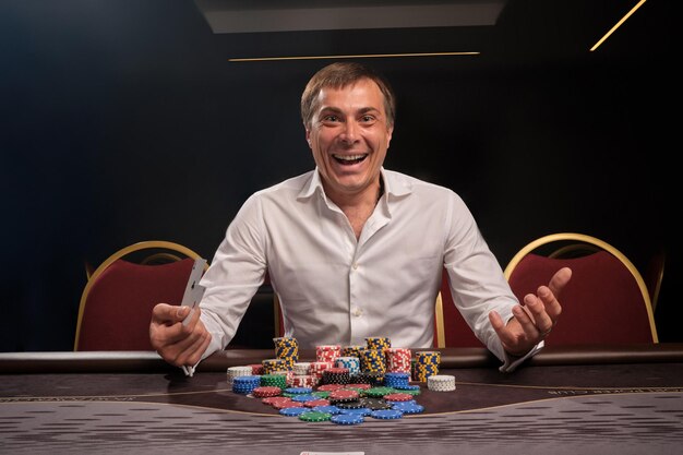 Un joven sonriente con una camisa clásica blanca está jugando al póquer sentado en la mesa del casino. Él se regocija en ganar y mirando a la cámara. Juegos de azar por dinero. Juegos de fortuna.