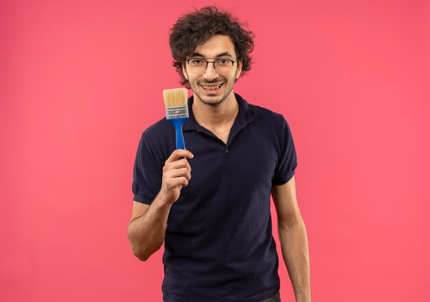 Joven sonriente en camisa negra con gafas ópticas sostiene cepillo y parece aislado en la pared rosa