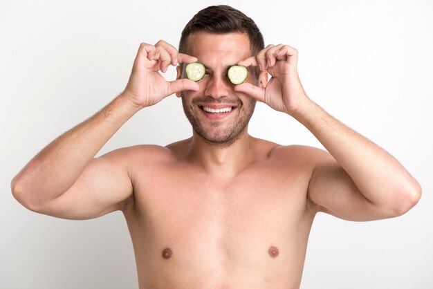 Joven sonriente sin camisa que cubre los ojos con una rodaja de pepino de pie contra la pared blanca