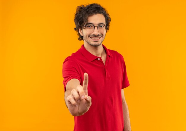 Joven sonriente en camisa roja con gafas ópticas gestos uno aislado en la pared naranja