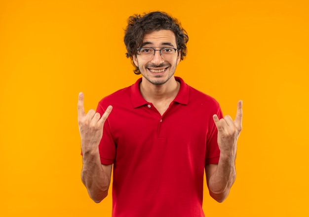 Joven sonriente en camisa roja con gafas ópticas gestos cuernos con las manos aisladas en la pared naranja