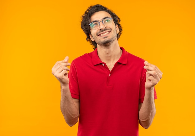 Joven sonriente en camisa roja con gafas ópticas gestos dinero con las manos aisladas en la pared naranja