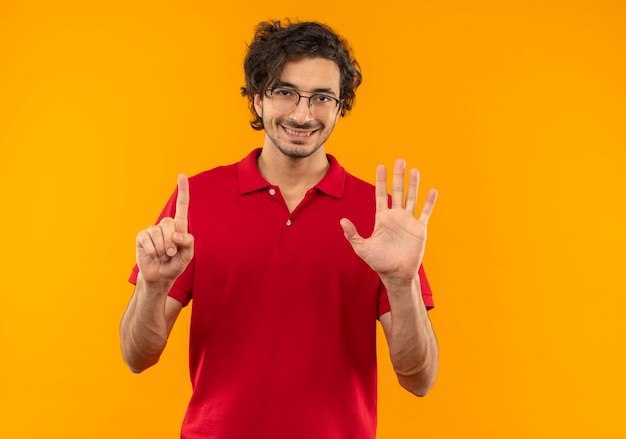 Joven sonriente en camisa roja con gafas ópticas gestos seis con los dedos aislados en la pared naranja