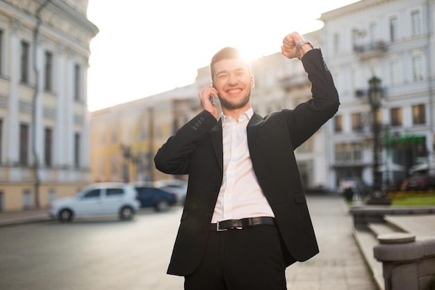 Joven sonriente con chaqueta negra y camisa blanca felizmente levantando la mano mientras habla por teléfono celular con una hermosa vista de la ciudad en el fondo