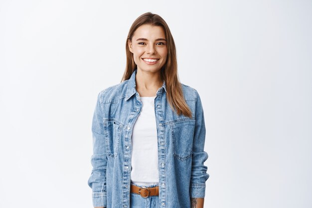 Joven sonriente a chica rubia con dientes perfectos mirando al frente, de pie relajado contra la pared blanca en ropa casual, estilo de vida y concepto de emociones