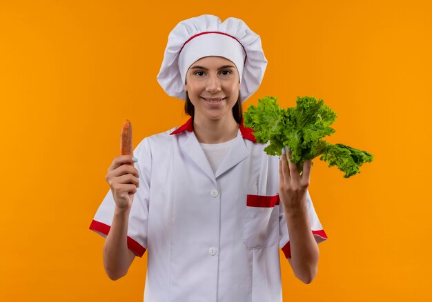 Joven sonriente cocinera caucásica en uniforme de chef sostiene zanahoria y ensalada aislado en espacio naranja con espacio de copia