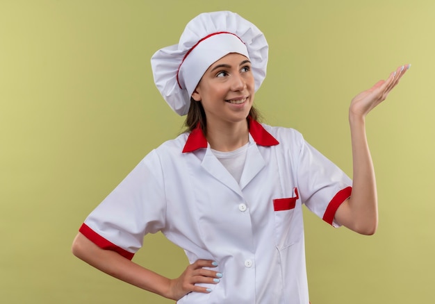 Joven sonriente cocinera caucásica en uniforme de chef se ve y apunta al lado en verde con espacio de copia