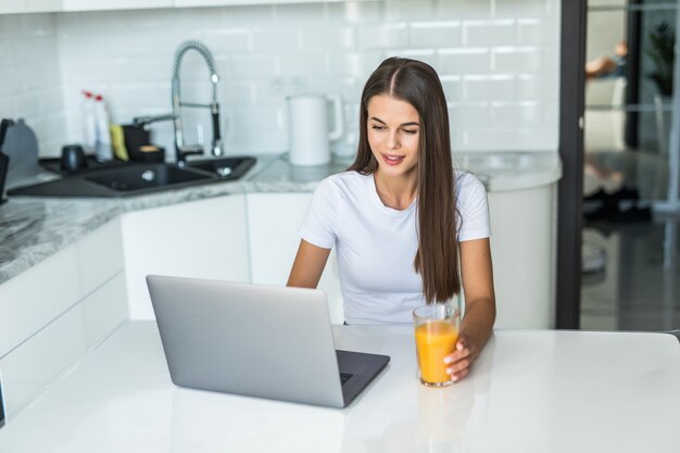 Joven sonriente desayunando en la cocina conectando con una computadora portátil y bebiendo un jugo de naranja saludable