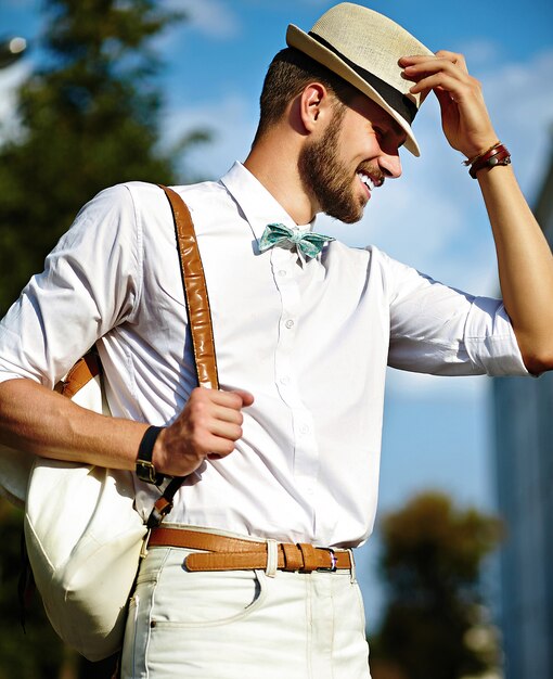 Joven sonriente elegante sexy modelo guapo hombre turista en estilo de vida casual de tela en la calle con sombrero con bolsa