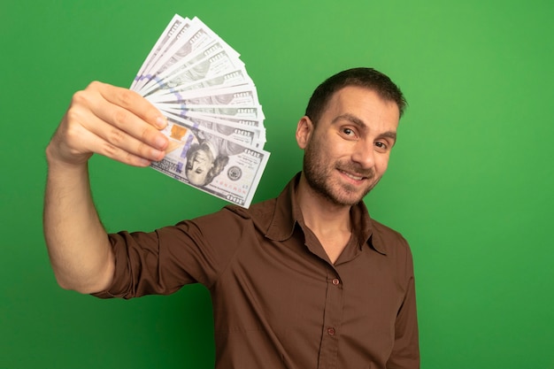 Joven sonriente estirando dinero hacia el frente mirando a la cámara aislada en la pared verde